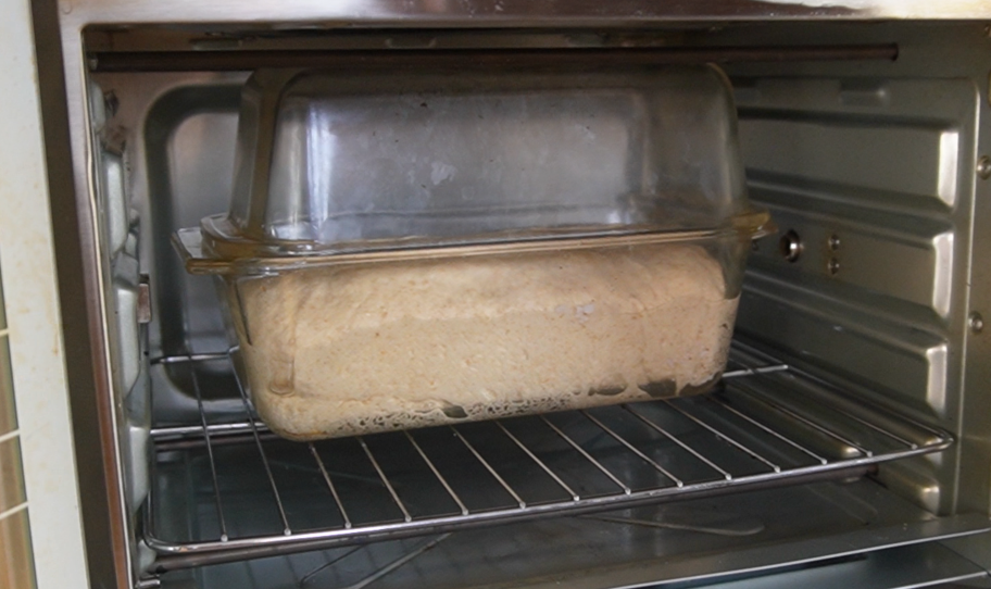 Bread dough inside a glass baking pan with a second empty glass baking pan placed on top upside down. This is inside an oven.